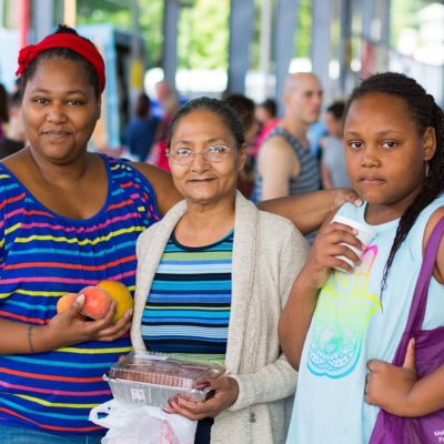 A group of three Black women holding fruit and a pie at a farmers market