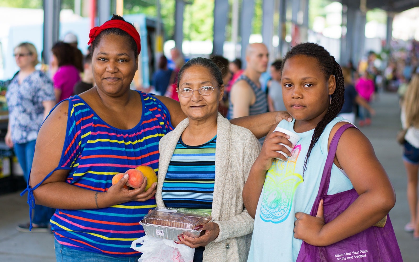 A group of three Black women holding fruit and a pie at a farmers market